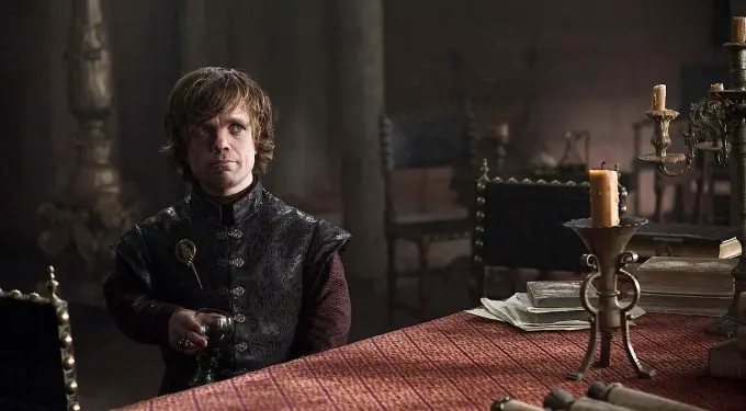 Tyrion Lannister seated at table holding a goblet in dimly lit room