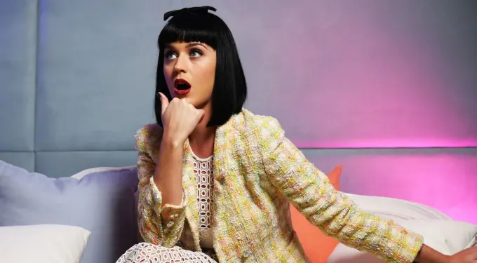 Katy Perry in colorful jacket poses sitting with surprised expression