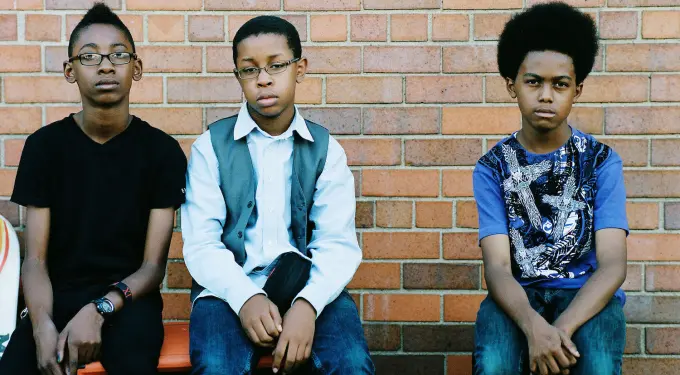 Three teenage boys sitting against brick wall, looking serious and still