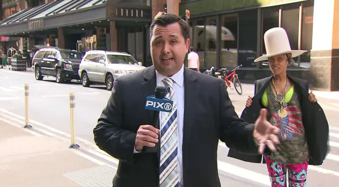 Reporter holding PIX11 microphone with woman in large white hat behind him
