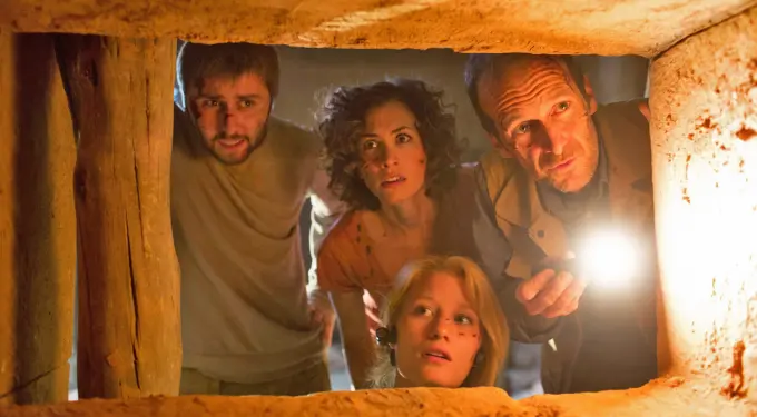 Four people with dirtied faces look into a stone opening, one holding flashlight