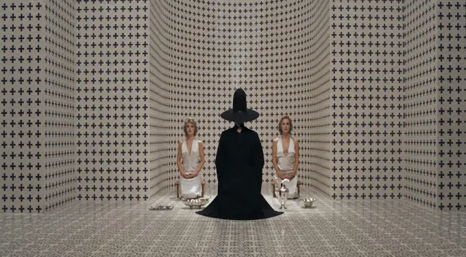 Two women kneel facing person in black robe and tall hat in patterned room