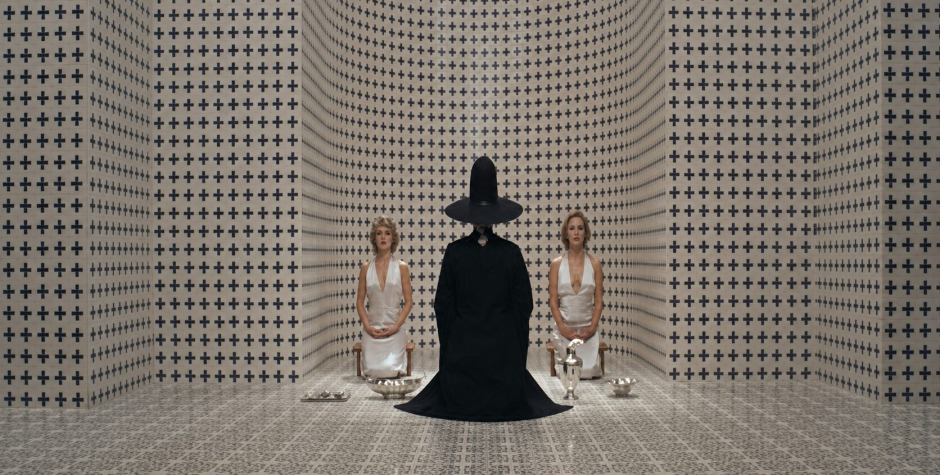 Two women kneel facing person in black robe and tall hat in patterned room
