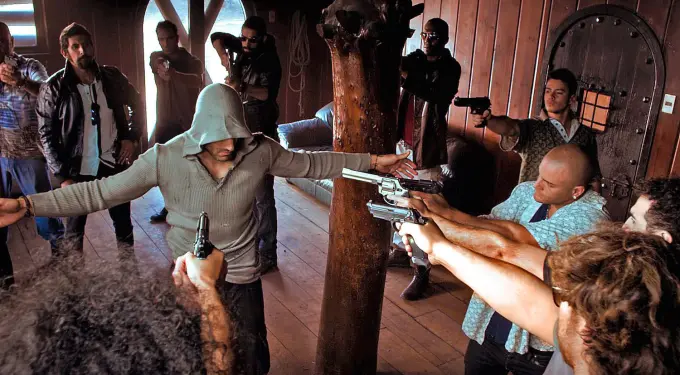 Man in hooded sweater held at gunpoint by multiple armed men indoors