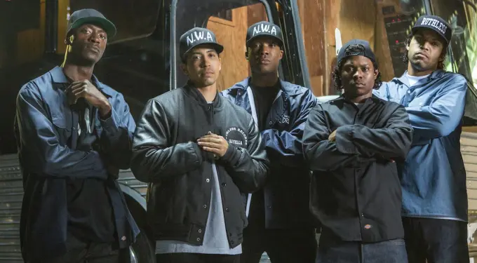 Group of five men in 90s hip-hop style clothes pose in front of bus