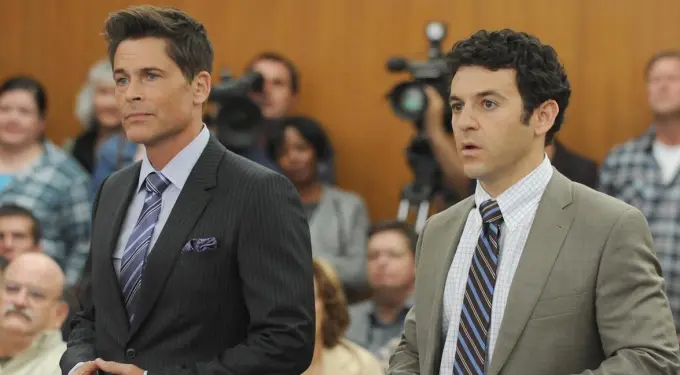 Two men in suits stand in courtroom with audience behind