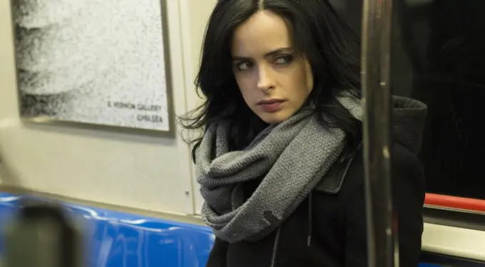 Jessica Jones on subway, wearing black coat and gray scarf, looking sideways