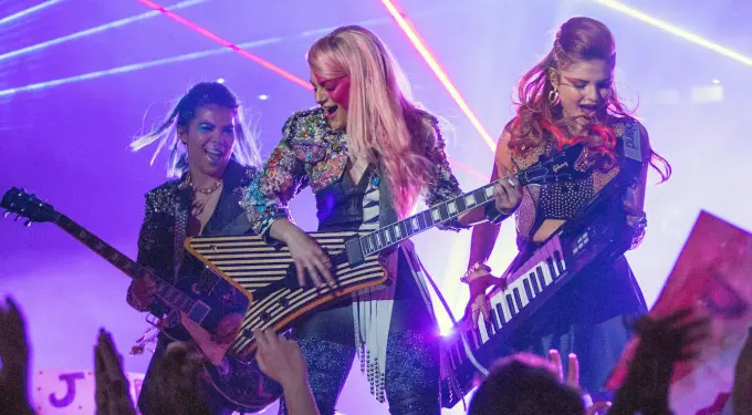 Three women play electric guitar and keytar on stage amid purple lighting