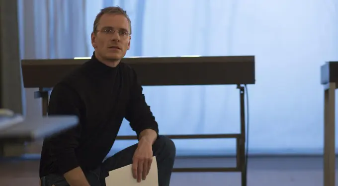 Michael Fassbender as Steve Jobs kneels holding papers by desk