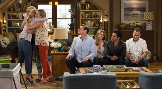 Two women hug, four others sit in living room on Fuller House set