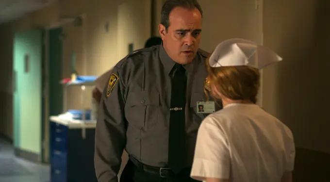 David Zayas in security guard uniform talks to nurse in hospital hallway
