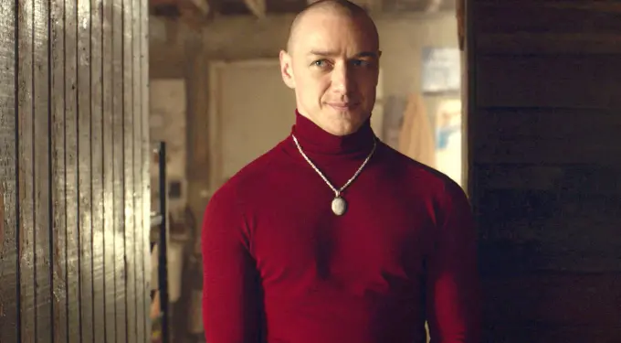 Man in red turtleneck and necklace standing indoors near wood paneling