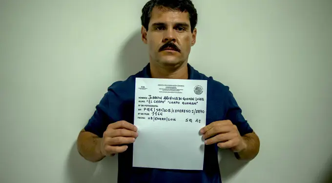 Man with mustache holding mugshot sign against a plain wall