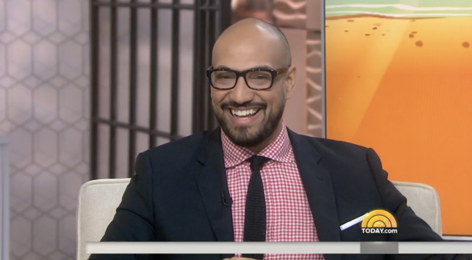 Man in glasses and suit smiling in a TV interview