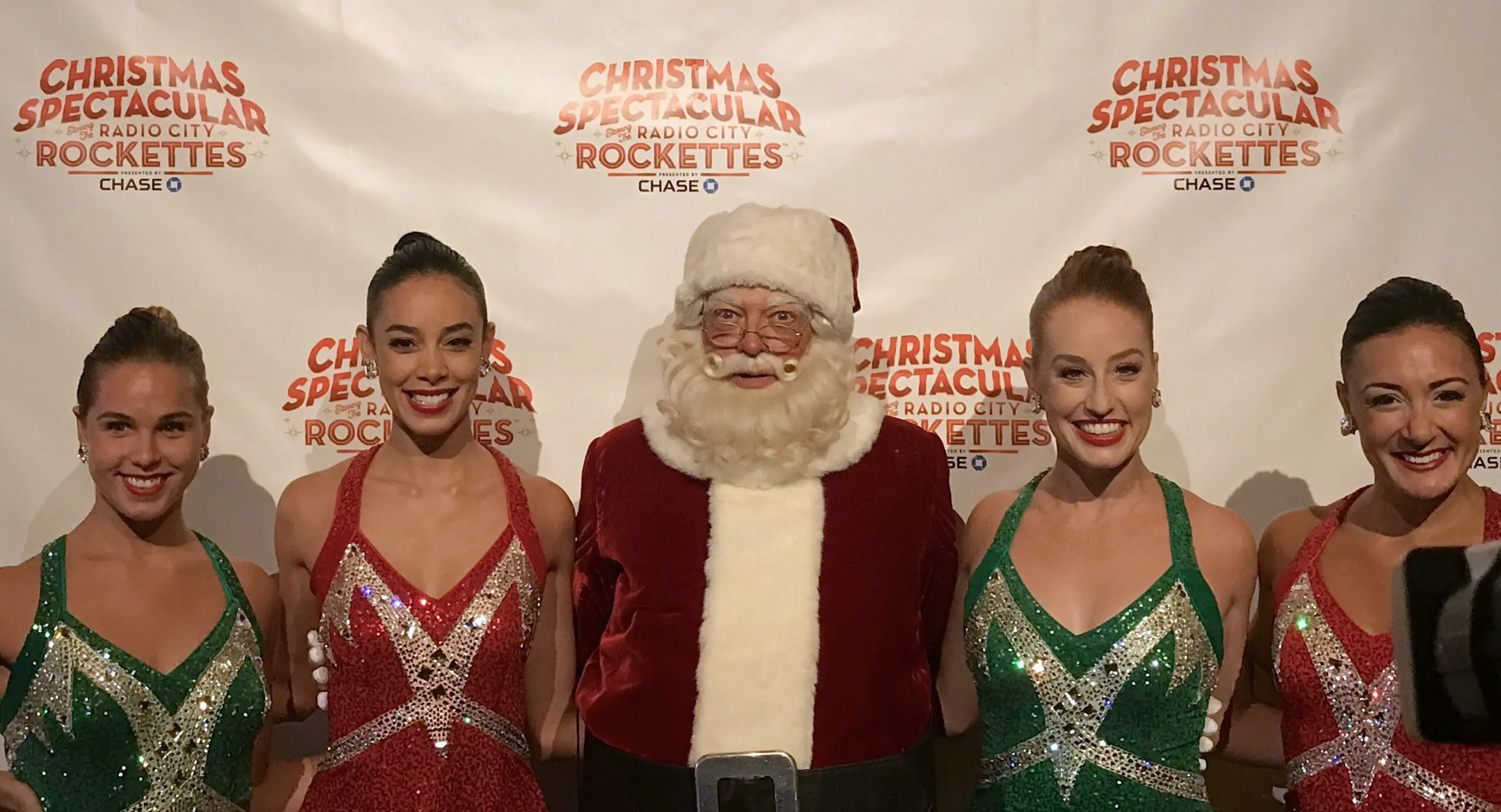Santa Claus stands with four Rockettes dancers in green and red costumes