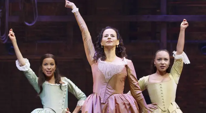Three women in period dresses with raised arms on stage
