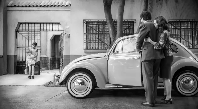Two adults near vintage car, woman embraces man; child and woman stand at gate