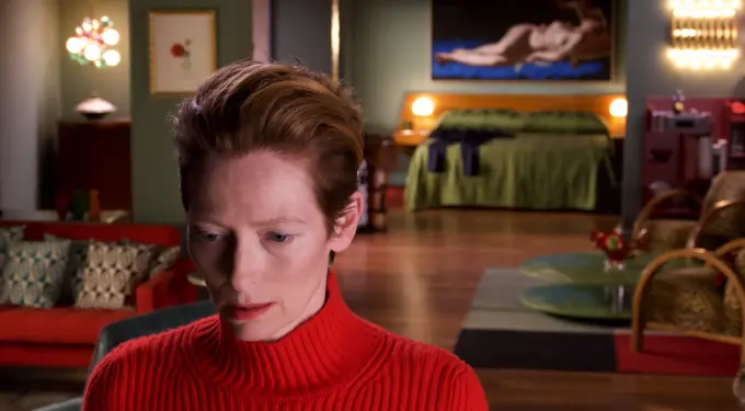 Tilda Swinton in red sweater looking down in moody room