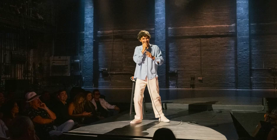 stand-up comedian holding microphone, performing to seated audience on dark stage